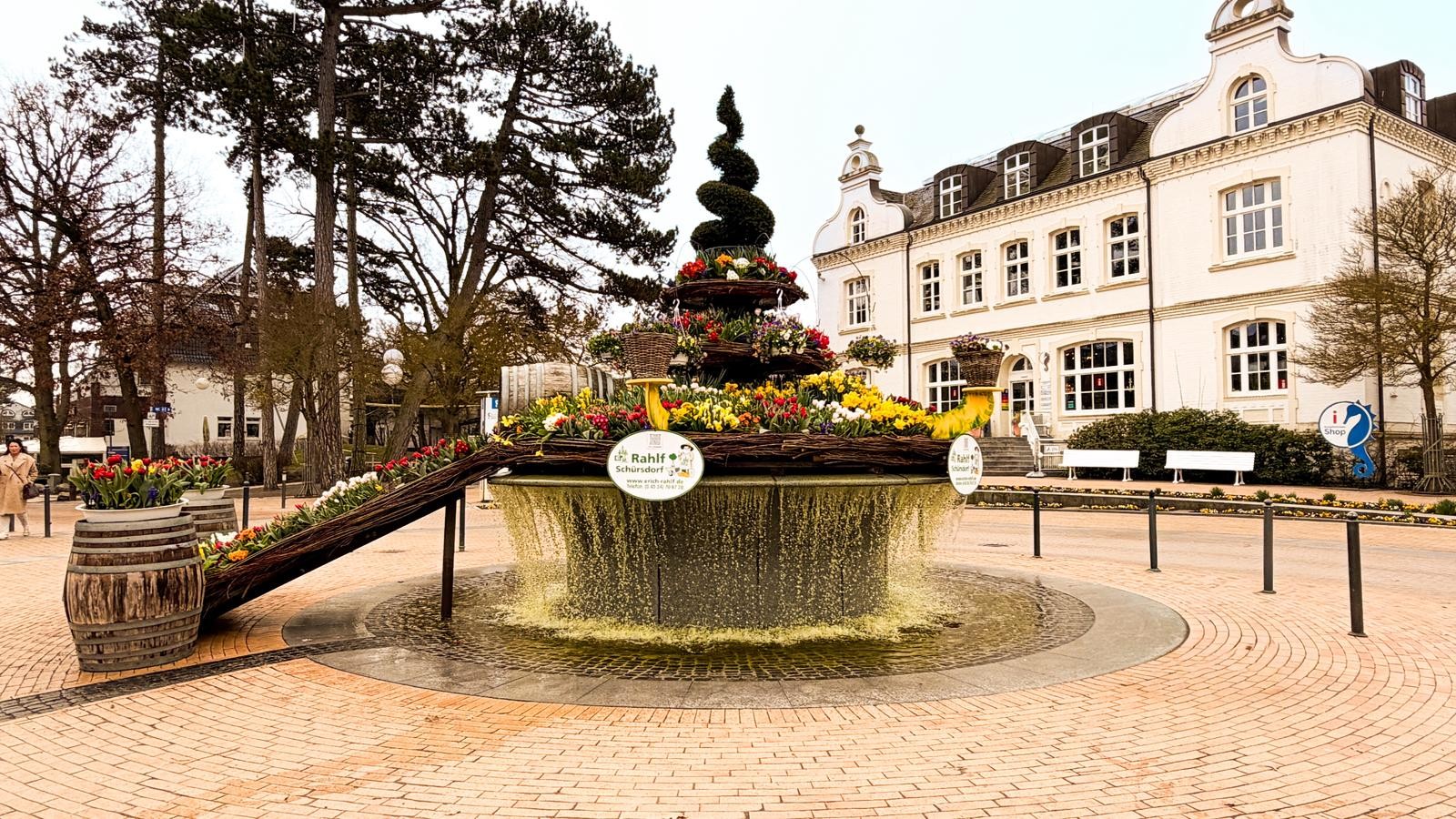 ein runder Brunnen mit fließendem Wasser und einer bepflanzten Blumeninsel der in der Mitte steht auf einem gepflasterten Platz.