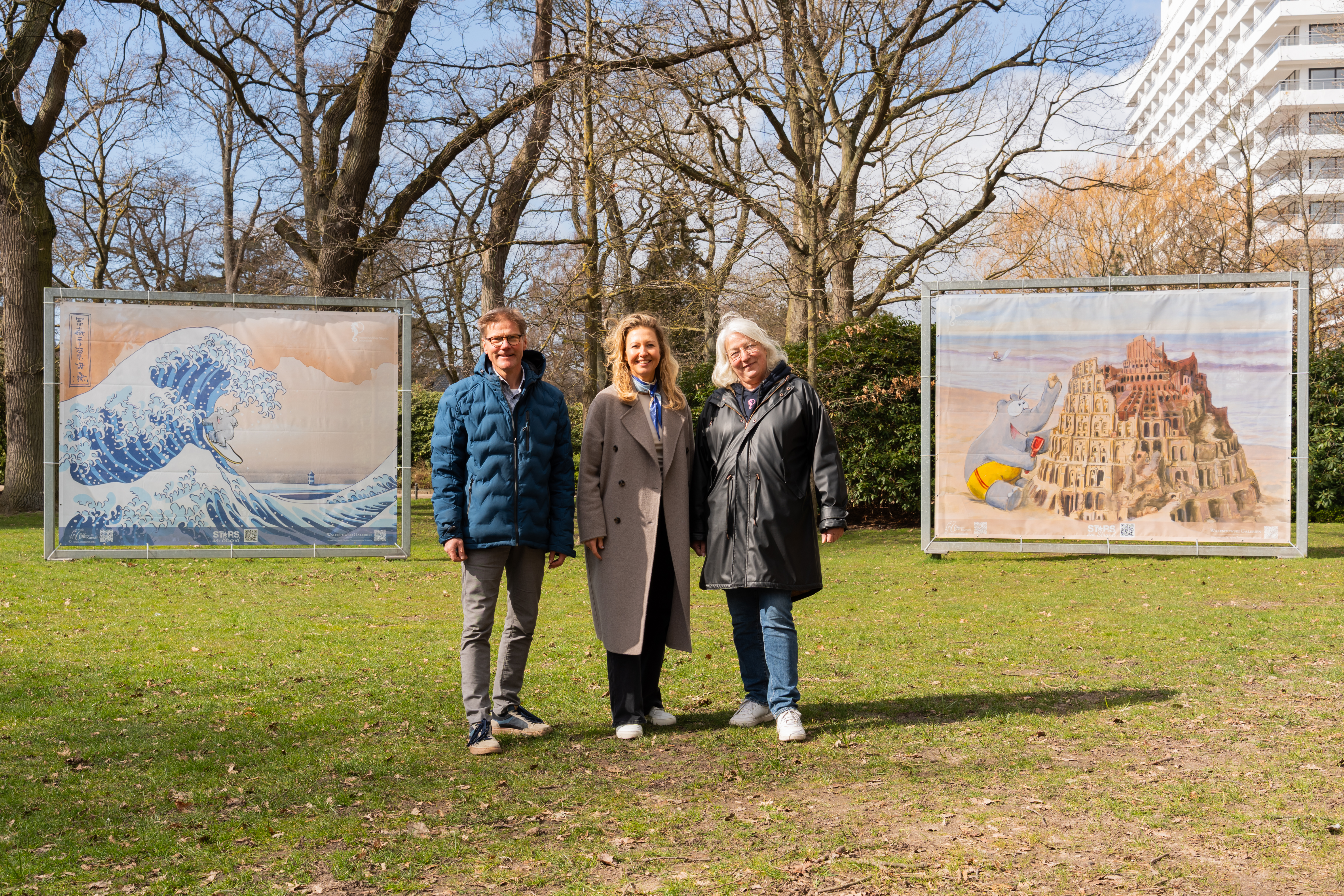 drei Personen auf einer Wiese in einem Park lächeln in die Kamera. Hinter ihnen sind zwei große gerahmte Kunstwerke ausgestellt. Links eine Darstellung einer großen Welle und rechts ein Bild eines turmartigen Gebäudes