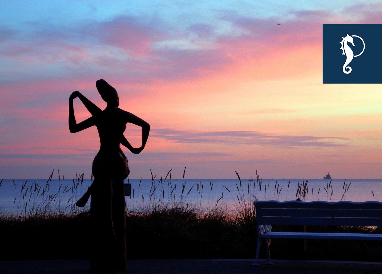 Skulptur einer sitzenden Frau mit einem Handtuch in der Abenddämmerung. Der Himmel leuchtet rosa, blau und gelb.