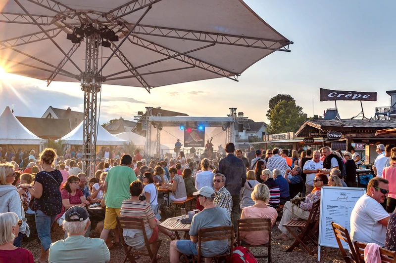 Große Veranstaltung in der Nachmittagssonne. Viele Menschen sitzen an Tischen und auf einer Bühne spielt eine Band.