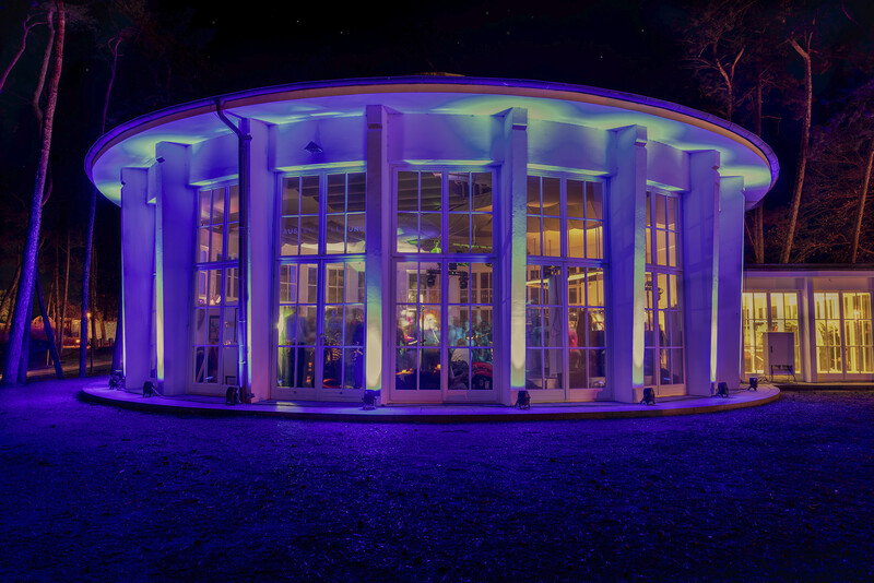 Rundes Gebäude mit großen Glasfenstern, nachts mit bunten Lichtern, durch die Fenster sind Menschen im Inneren zu sehen bei einer Veranstaltung