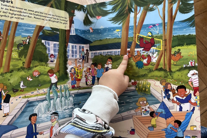 Eine Kinderhand zeigt auf eine Figur im Wimmelbuch.