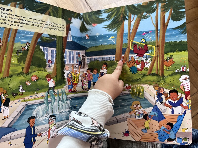 Eine Kinderhand zeigt auf eine Figur im Wimmelbuch. 
