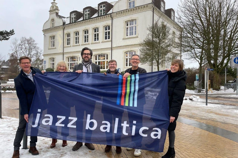 Sechs Menschen mit einem Banner mit dem Schriftzug Jazz Baltica in der Hand vor einem alten Gebäude