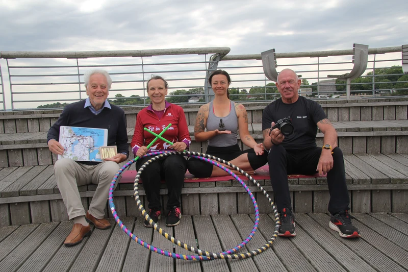 Vier Personen sitzen auf einer Bank am Meer und halten verschiedene Sachen in den Händen wie einen Fotoapperat und Hula-Hoop Reifen.