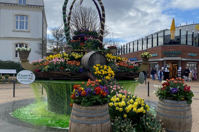 Bunt bepflanzter Osterbrunnen und im Hintergrund das alte Rathaus von Timmendorfer Strand