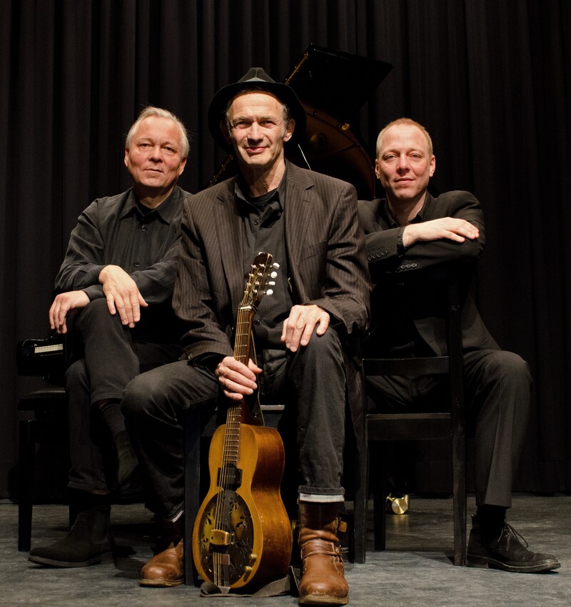 Drei Musiker sitzen und schauen in die Kamera, der Musiker im Vordergrund hält eine Gitarre in der Hand.