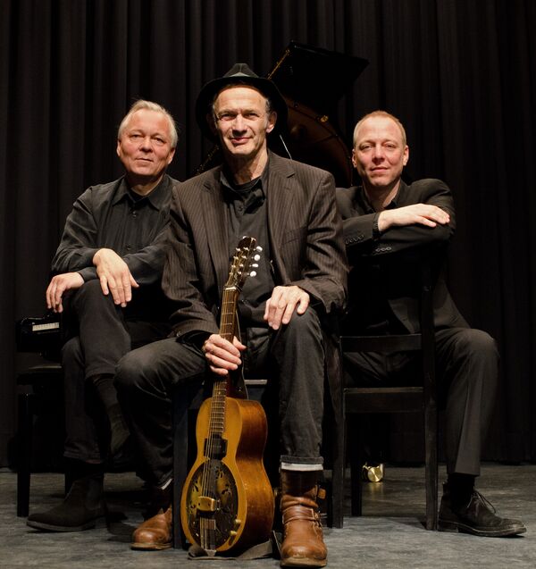 Drei Musiker sitzen und schauen in die Kamera, der Musiker im Vordergrund hält eine Gitarre in der Hand.