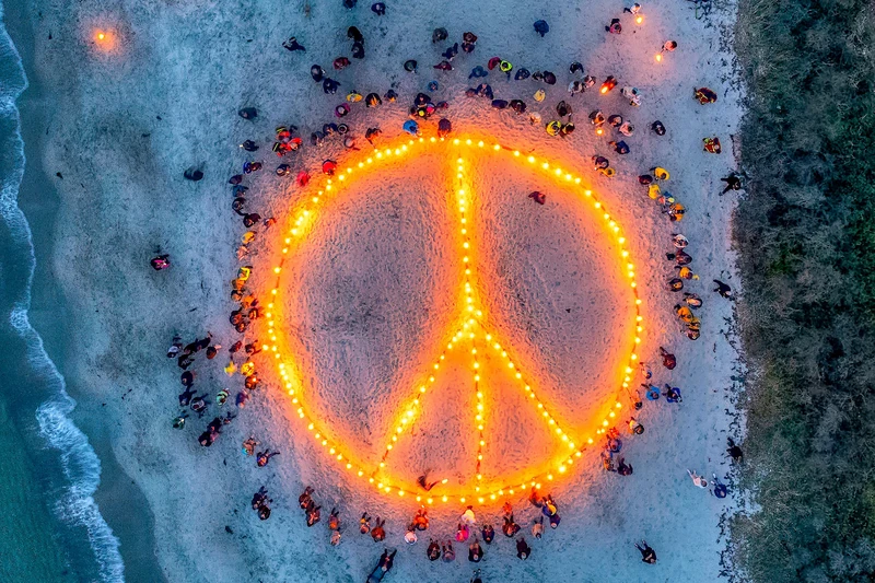 Luftaufnahme: Gigantisches Friedenssymbol aus Feuer im Sand am Strand, viele Menschen stehen darum herum