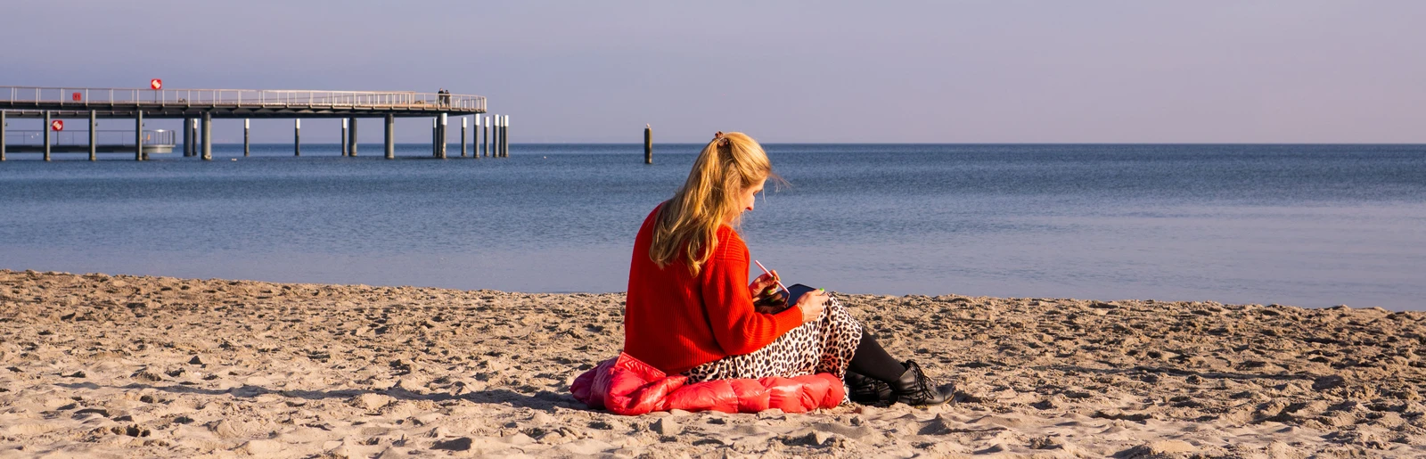 Lara Klewin sitzt im Sand am Strand mit einem Ipad in der Hand und arbeitet. Der Himmel ist blau und die Seebrücke ist zu sehen.