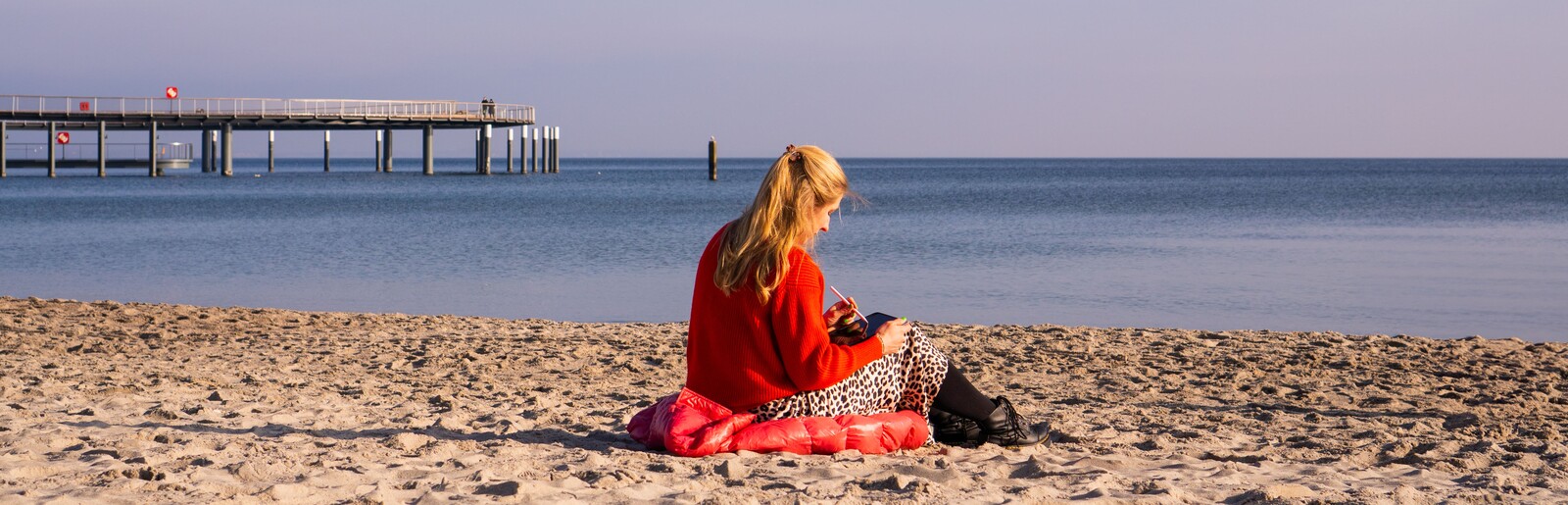 Lara Klewin sitzt im Sand am Strand mit einem Ipad in der Hand und arbeitet. Der Himmel ist blau und die Seebrücke ist zu sehen. 