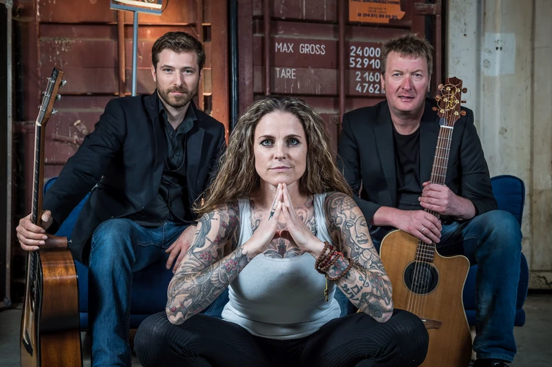 Bandfoto. Eine Frau im Vordergrund und zwei Männer mit Instrumenten hinter ihr.