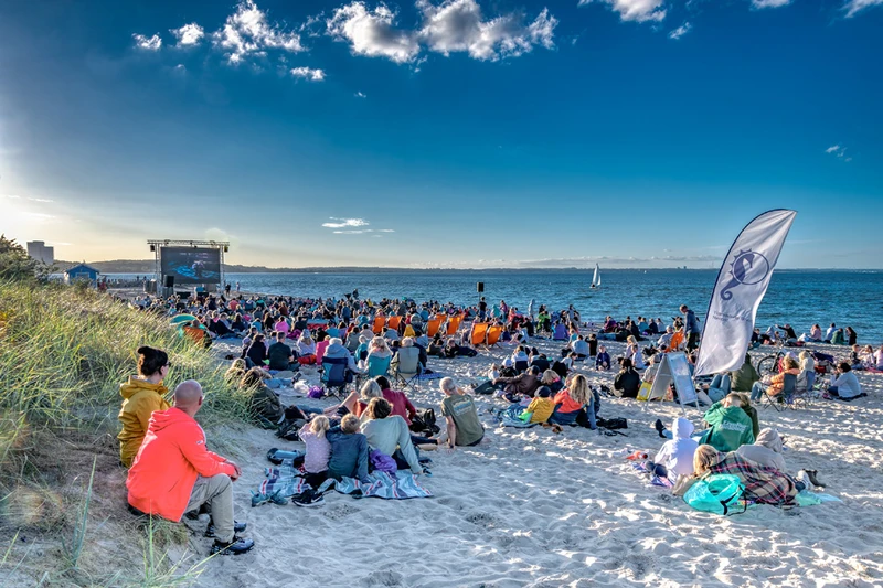 Viele Menschen sitzen am Strand vor einer großen aufgebauten Leinwand und schauen einen Film