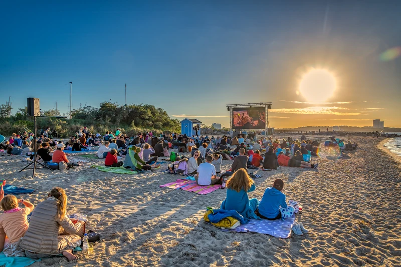 Foto am Strand beim Sonnenuntergang. Im Vordergrund sitzen Menschen und schauen auf einer großen Leinwand einen Film 