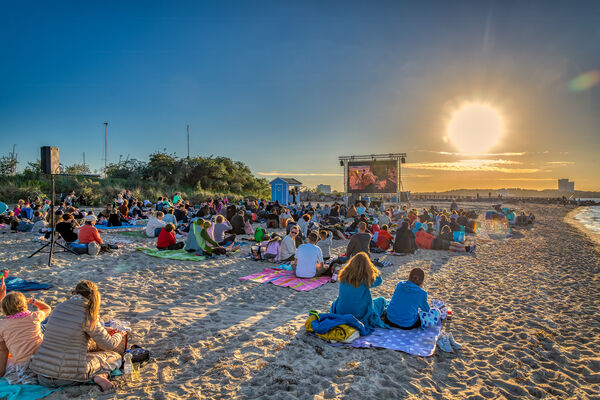 Foto am Strand beim Sonnenuntergang. Im Vordergrund sitzen Menschen und schauen auf einer großen Leinwand einen Film