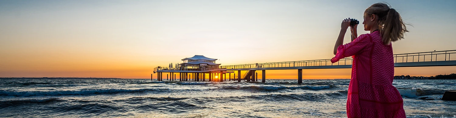 Frau mit Fernglas steht beim Sonnenuntergang am Strand und guckt auf das Teehaus