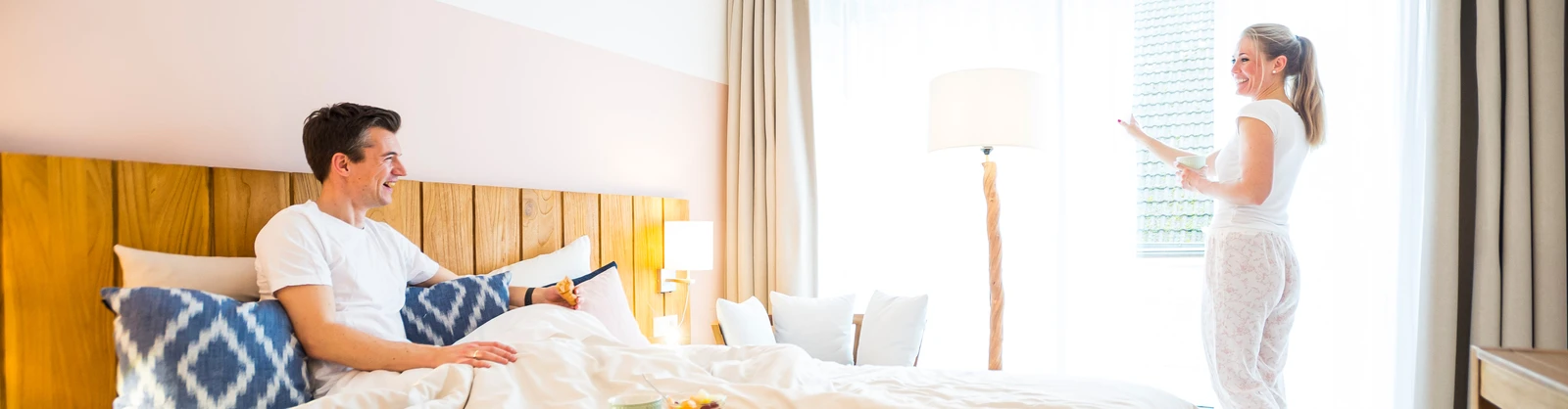 Ein Pärchen in einem Hotelzimmen. Er im Bett und sie am Fenster stehend.