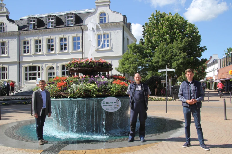 Drei Personen stehen vor dem zu Pfingsten geschmückten Stadtbrunnen von Timmendorf