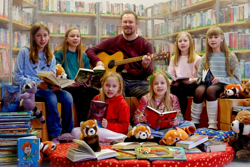 Sechs Kinder in einer Bücherei umgeben von Stapeln mit Büchern. In der Mitte ein Mann mit einer Gitarre.