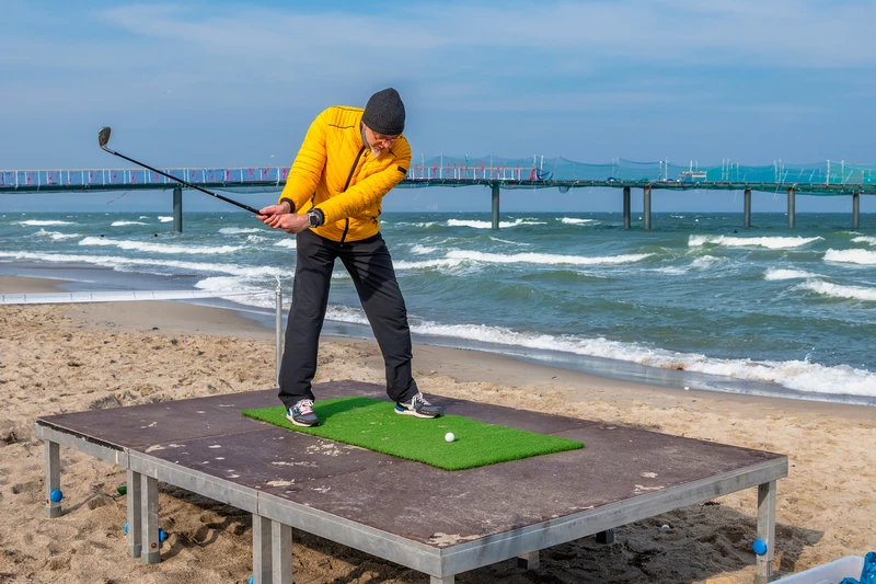 Mann steht auf einer Plattform am Strand mit Schläger und Golfball und bereitet den Abschlag vor