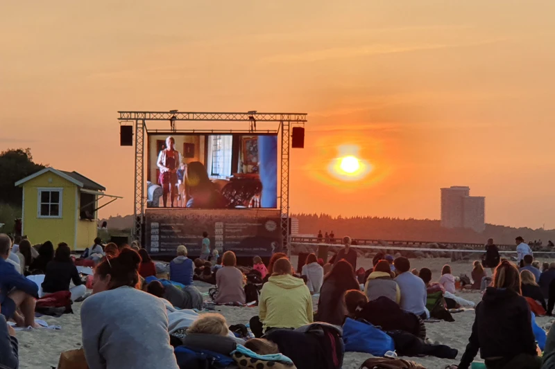 Viele Menschen sitzen am Strand in der Abendsonne und schauen einen Film auf einer großen Leinwand
