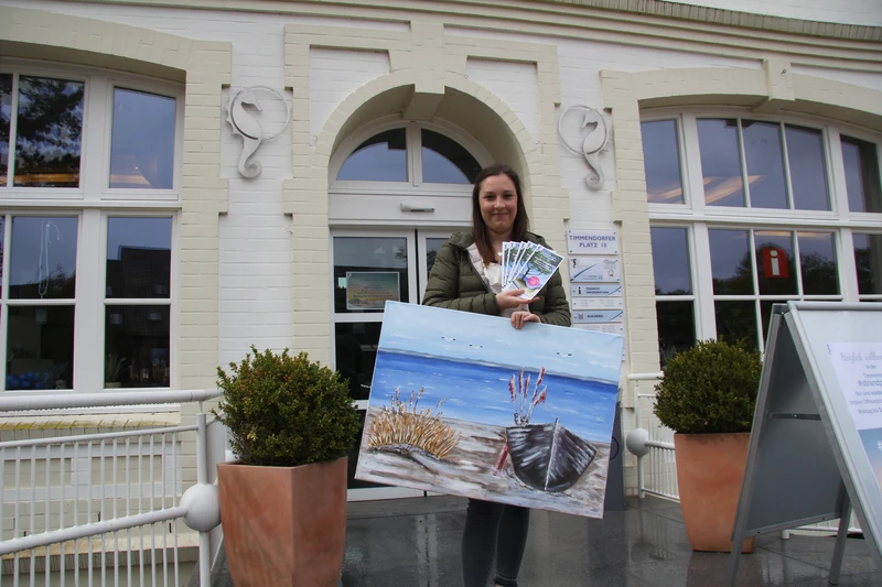 Frau steht mit einem großen Bild von einem Ruderboot und einer Hand voll Flyer draußen vor einer Eingangstür