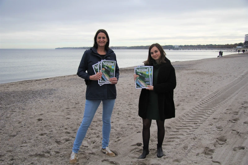 Zwei Frauen mit jeweils einem Stapel Strandmagazine in den Händen am Strand