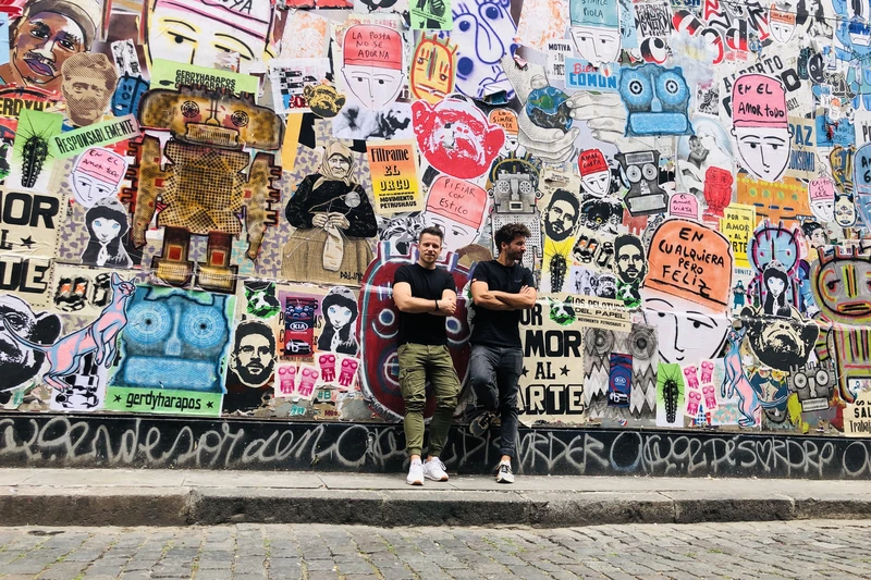 Zwei Männer angelehnt an eine Wand voller Plakate und Graffiti.