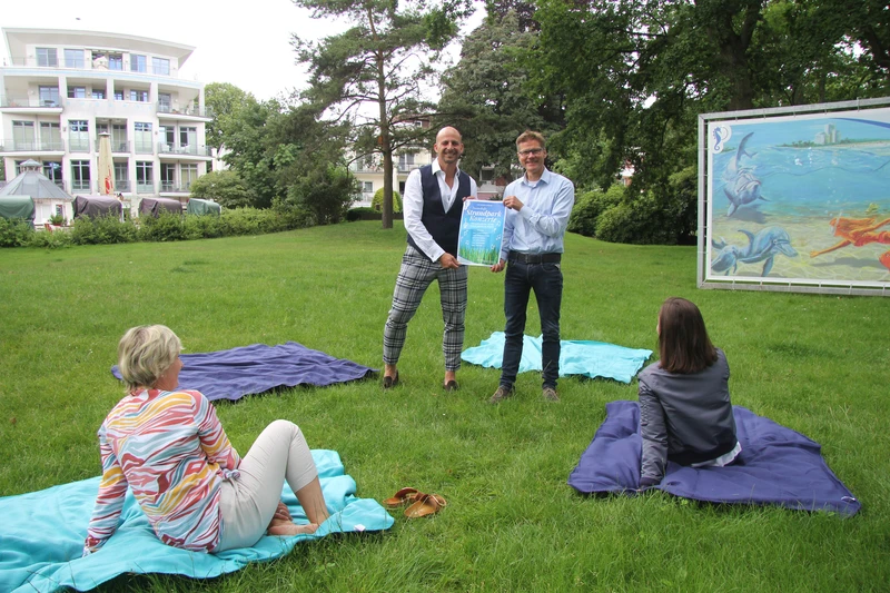 Einladung zu Strandpark-Konzerten: zwei Männer halten Plakat hoch, zwei Frauen sitzen auf Picknickdecken im Gras