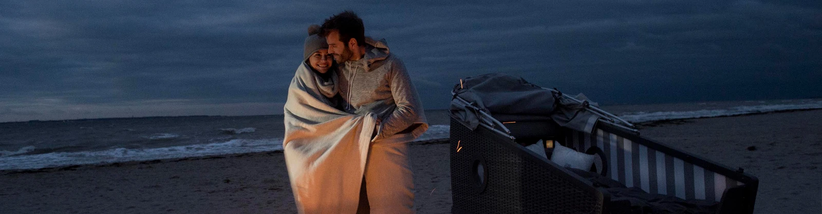 Pärchen in eine Decke gehüllt steht am Strand vor einem Feuerkorb und einem Schlafstrandkorb.