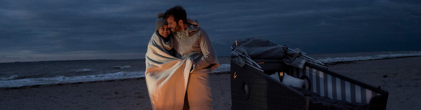 Pärchen in eine Decke gehüllt steht am Strand vor einem Feuerkorb und einem Schlafstrandkorb.