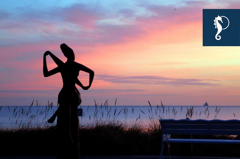 Skulptur einer sitzenden Frau mit einem Handtuch in der Abenddämmerung. Der Himmel leuchtet rosa, blau und gelb.