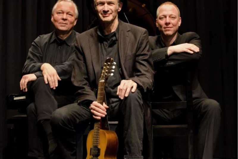 Bandfoto der drei Männer mit schwarzem Hintergrund. Sie tragen alle Anzüge und der Mann in der Mitte hält eine Mandoline.