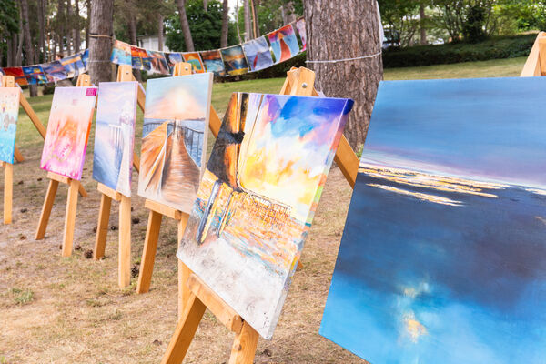 Mehrere bunte Gemälde auf Staffeleien stehen im Freien auf sandigen Boden, im Hintergrund hängen weitere Bilder zwischen Bäumen