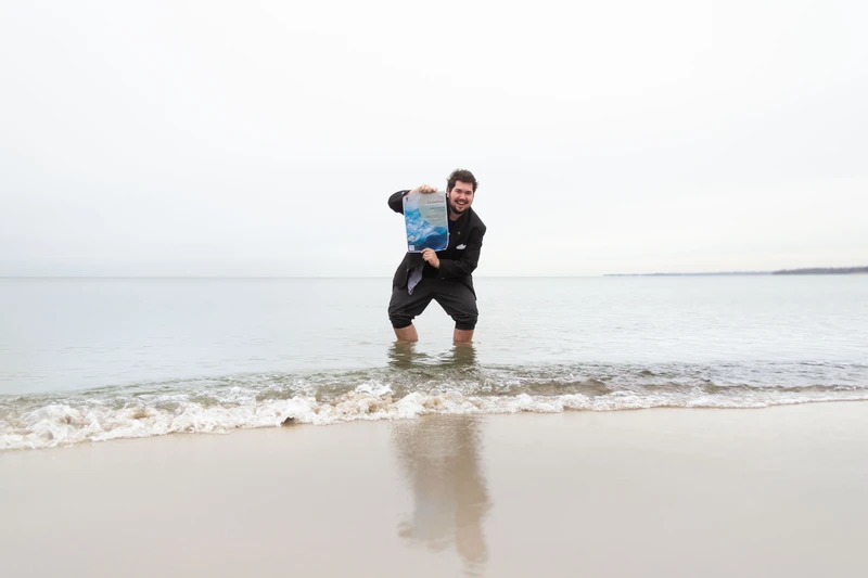Der Auszubildende Melvin Kamp mit dem Plakat für den Kunstwettbewerb 2024 in der Hand steht mit hochgekrempelter Hose in der Ostsee