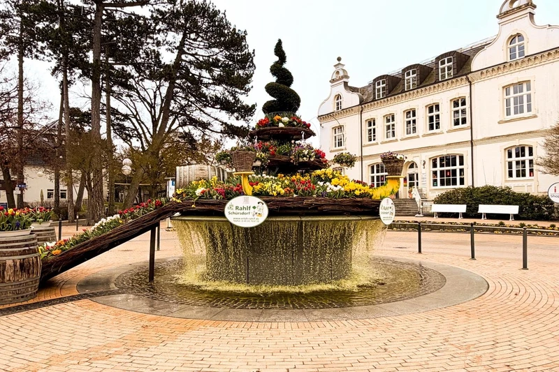 ein runder Brunnen mit fließendem Wasser und einer bepflanzten Blumeninsel der in der Mitte steht auf einem gepflasterten Platz. 