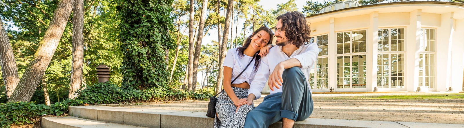Pärchen sitzt auf einer Treppe vor einem gläsernden Gebäude, der Trinkkurhalle.