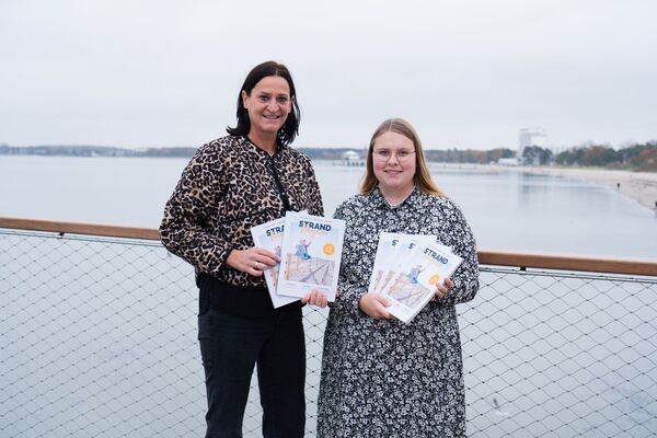 Marketingleiterin und Projekt- und Kampagnenmanagerin freuen sich auf der neuen Maritim Seebrücke über das Strandmagazin 2025