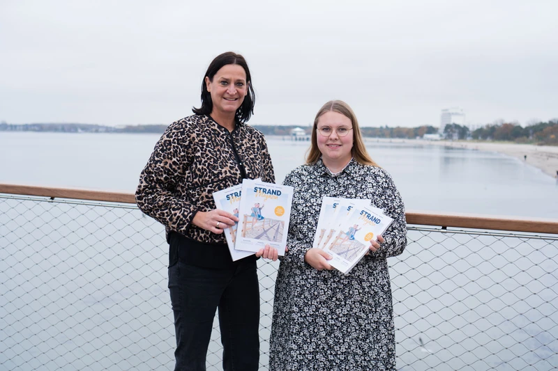 Marketingleiterin und Projekt- und Kampagnenmanagerin freuen sich auf der neuen Maritim Seebrücke über das Strandmagazin 2025