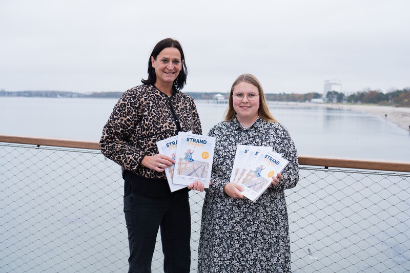 Marketingleiterin und Projekt- und Kampagnenmanagerin freuen sich auf der neuen Maritim Seebrücke über das Strandmagazin 2025