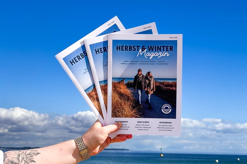 In der Hand werden die neuen Herbst-Winter Magazine gehalten, im Hintergrund sieht man die Ostsee und den blauen Himmel