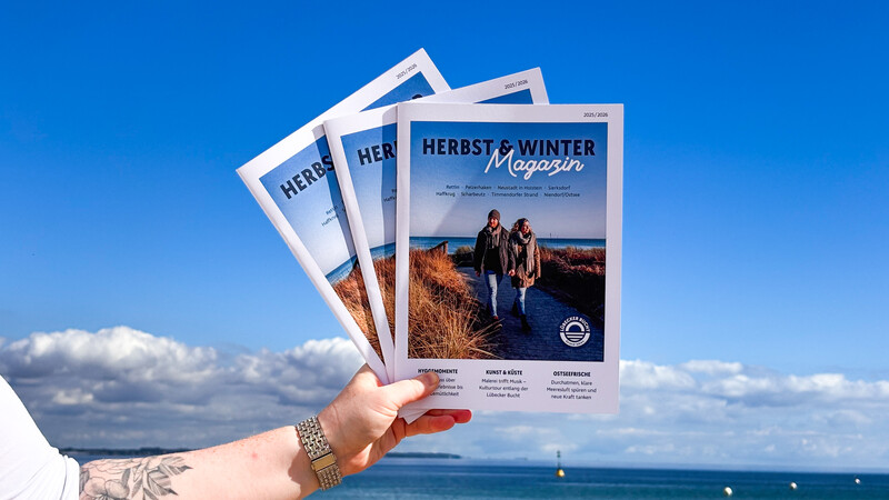 In der Hand werden die neuen Herbst-Winter Magazine gehalten, im Hintergrund sieht man die Ostsee und den blauen Himmel