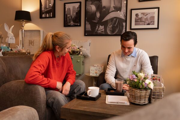 Ein Mann und eine Frau sitzen in einem gemütlichen Wohnzimmer auf einem Sofa und unterhalten sich. Der Mann zeigt etwas auf einem Tablet oder Papier. Auf dem Couchtisch stehen eine Tasse, eine Zeitung und eine Blumendekoration. Im Hintergrund hängen Schwarz-Weiß-Bilder, und eine Lampe sorgt für eine warme Atmosphäre.