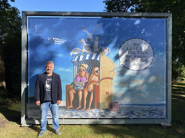 Cartoonist steht vor einer großen Plane in einem Stahlrahmen, auf der sein Cartoon aufgedruckt ist
