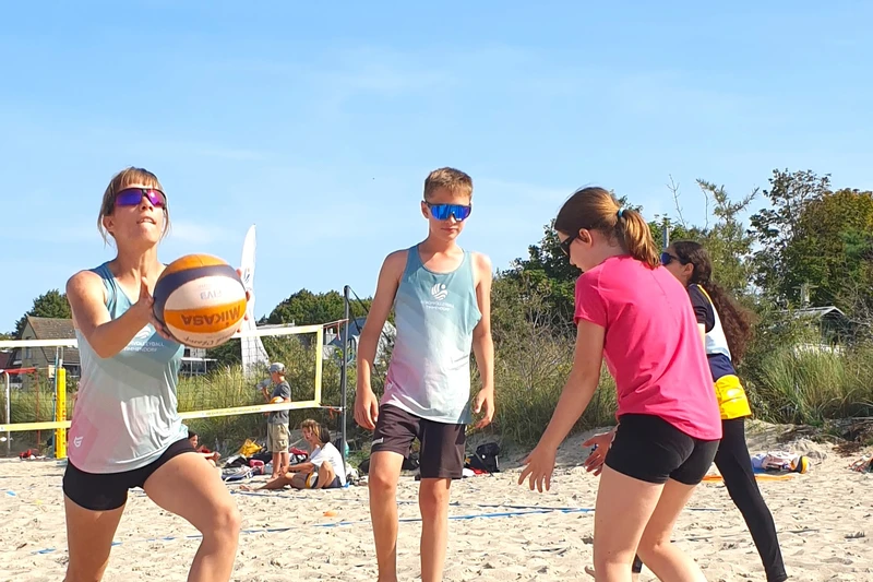 Drei Kinder die mit einem Volleyball spielen, einer hat den Ball unter seinem Fuß und die andere fängt grad an zu werfen 