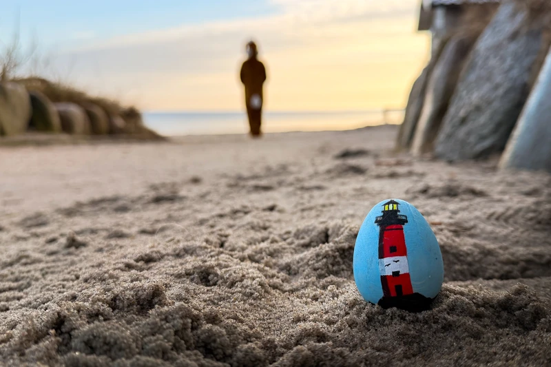 Ein bemaltes Ei mit Leuchtturm drauf im Vordergrund im Sand und dahinter ein Hase richtung Meer hoppeln