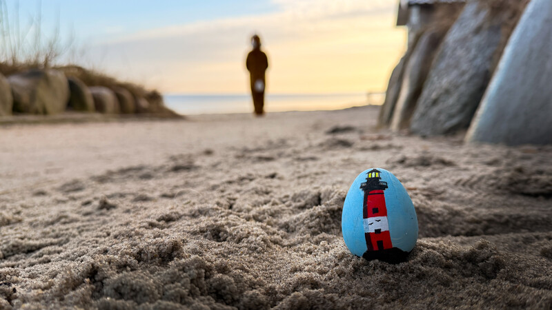 Ein bemaltes Ei mit Leuchtturm drauf im Vordergrund im Sand und dahinter ein Hase richtung Meer hoppeln