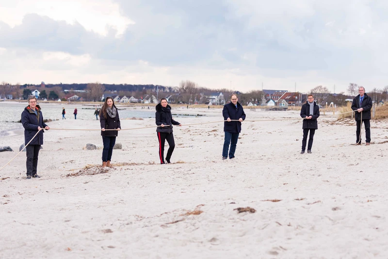 Sechs Personen stehen am Strand und halten ein Seil fest mit viel Abstand zueinander