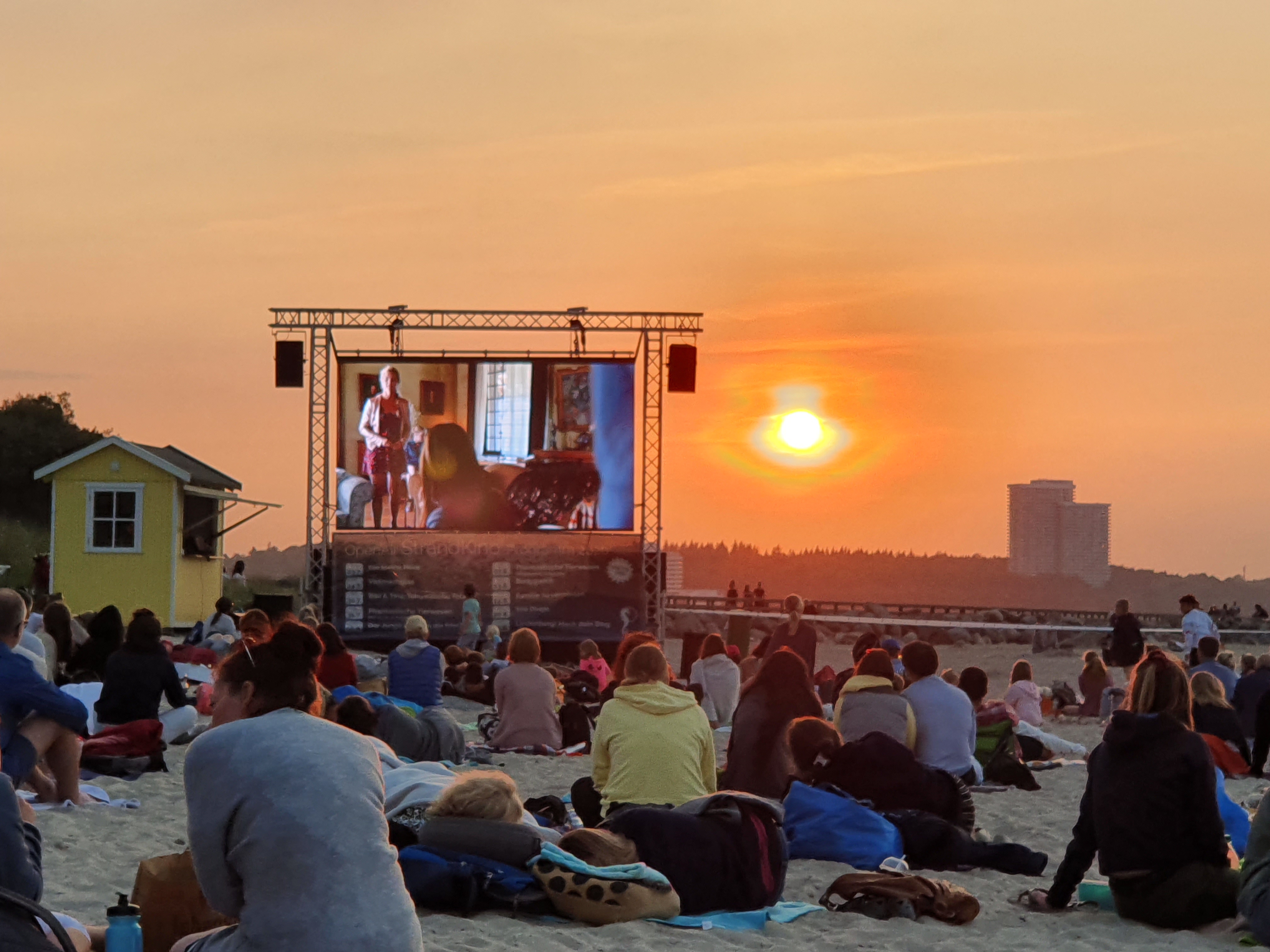 Viele Menschen sitzen am Strand in der Abendsonne und schauen einen Film auf einer großen Leinwand