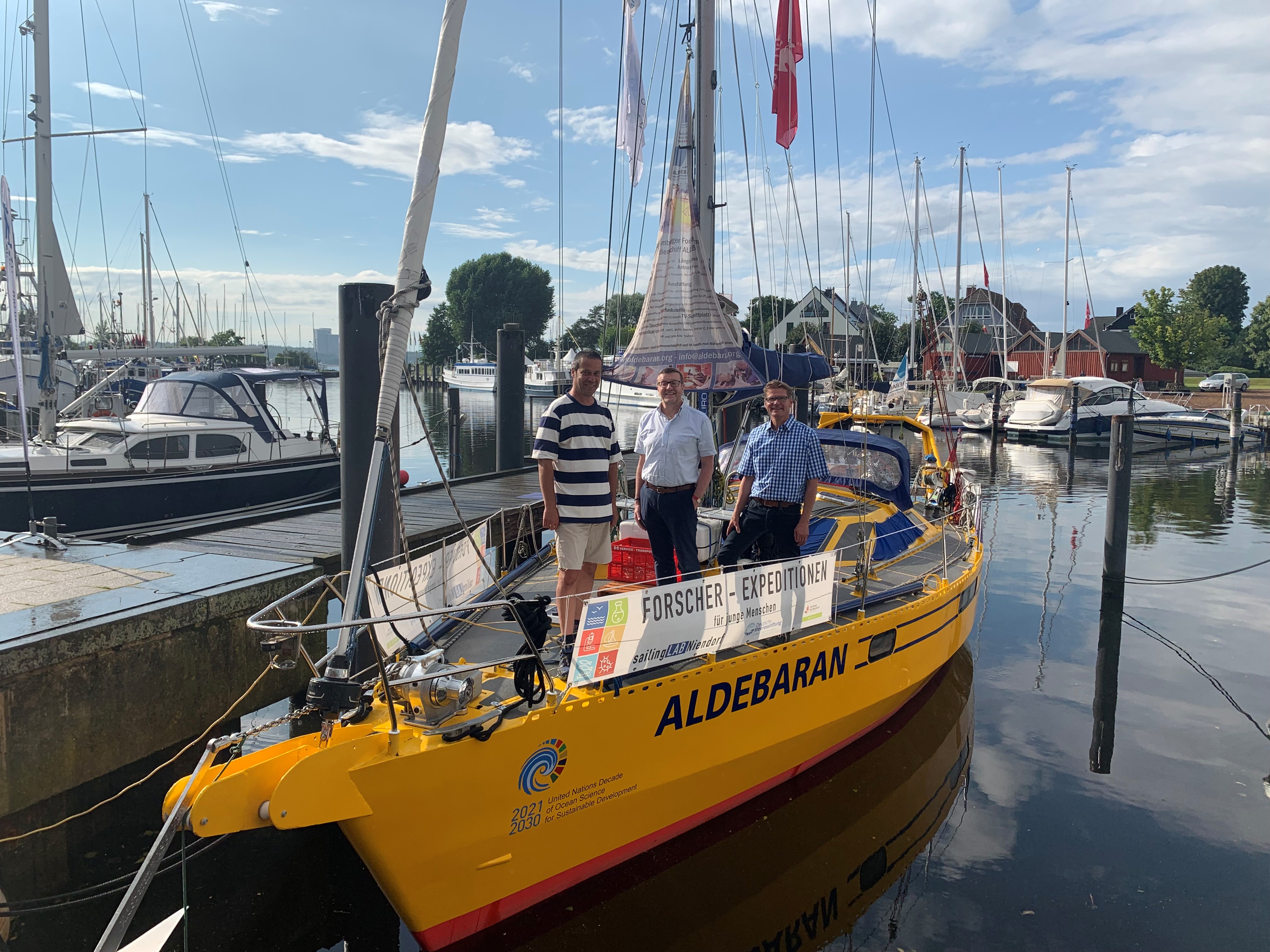 Das gelbe Forschungsschiff Aldebaran im Hafen mit drei Männern darauf